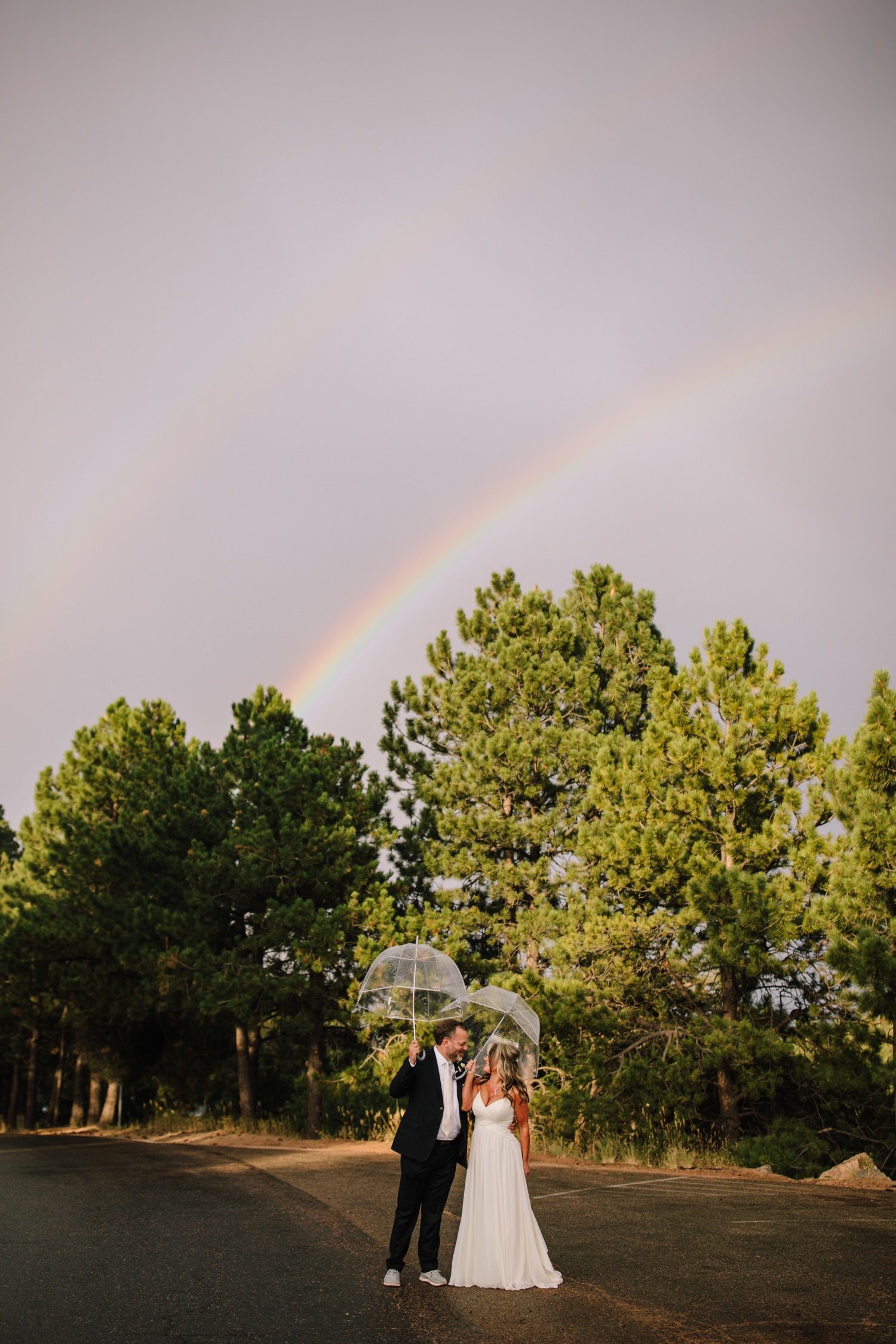 Bride and groom with double rainbow and clear umbrellas looking at each other