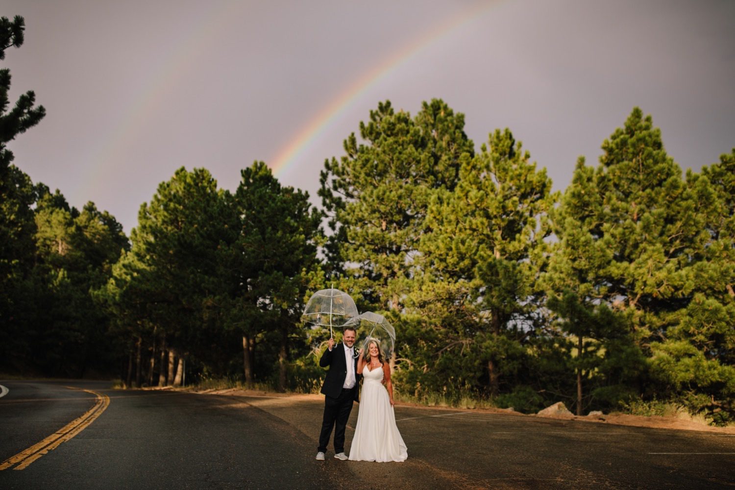 Bride and groom with double rainbow and clear umbrellas looking at the camera